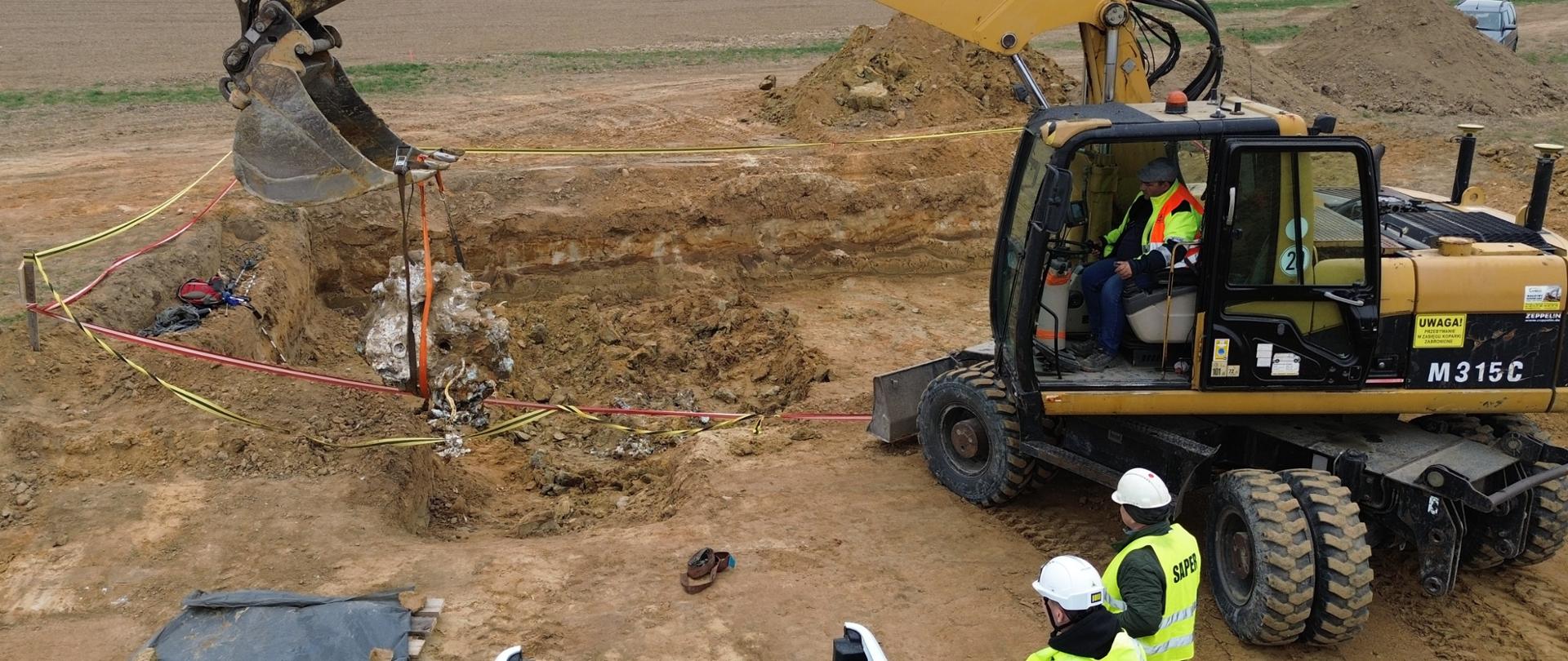 na zdjęciu widać koparkę, która wykopuje elementy samolotu z ziemi. Na zdjęciu widać sapera oraz przedstawiciela wykonawcy 