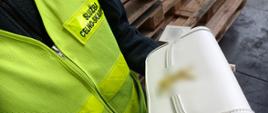 Osoba w kamizelce „Służba Celno‑Skarbowa” prezentuje jasną torebkę na tle drewnianych palet i betonowej podłogi.