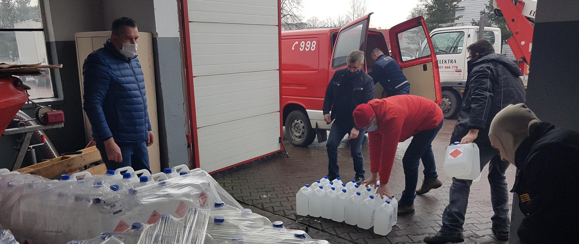 Na załączonym zdjęciu widać magazyn w którym na paletach typu EURO ustawione są pojemniki z płynem do dezynfekcji oraz pięciu druhów ochotniczych jednostek straży którzy przenoszą pojemniki z płynem do dezynfekcji i ładują na samochody typu bus . Po lewej stronie zdjęcia jest widoczny pracownik wydziału kwatermistrzowsko- finansowego Komendy Miejskiej PSP w Kaliszu który wydaje według rozdzielnika odpowiednie ilości płynów na poszczególne gminy powiatu kaliskiego. Wszystkie osoby maja na twarzy maseczki . Strażacy ochotniczych jednostek OSP będą rozwozić płyn oraz maseczki chirurgiczne do wszystkich jednostek ochotniczych straży pożarnych które są zaangażowane w przewóz osób do punktów szczepień na covid-19. 