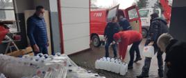 Na załączonym zdjęciu widać magazyn w którym na paletach typu EURO ustawione są pojemniki z płynem do dezynfekcji oraz pięciu druhów ochotniczych jednostek straży którzy przenoszą pojemniki z płynem do dezynfekcji i ładują na samochody typu bus . Po lewej stronie zdjęcia jest widoczny pracownik wydziału kwatermistrzowsko- finansowego Komendy Miejskiej PSP w Kaliszu który wydaje według rozdzielnika odpowiednie ilości płynów na poszczególne gminy powiatu kaliskiego. Wszystkie osoby maja na twarzy maseczki . Strażacy ochotniczych jednostek OSP będą rozwozić płyn oraz maseczki chirurgiczne do wszystkich jednostek ochotniczych straży pożarnych które są zaangażowane w przewóz osób do punktów szczepień na covid-19. 