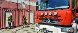 Samochód ratowniczo-gaśniczy na tle dwójki strażaków szykujących się do wejścia do budynku