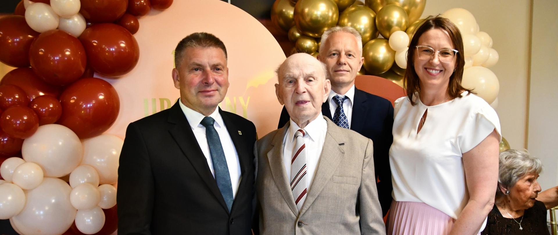 Trzech mężczyzn w garniturach i kobieta stoją na tle ścianki z balonami. W środku Jubilat. Wszyscy uśmiechają się do zdjęcia.