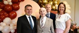 Trzech mężczyzn w garniturach i kobieta stoją na tle ścianki z balonami. W środku Jubilat. Wszyscy uśmiechają się do zdjęcia.