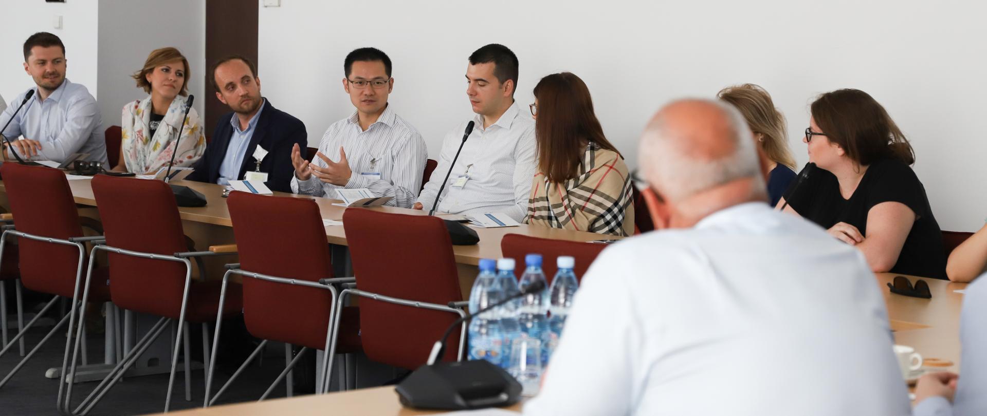 Widok zza placów wiceministra na gości siedzących za stołem. Jeden z nich mówi i gestykuluje.