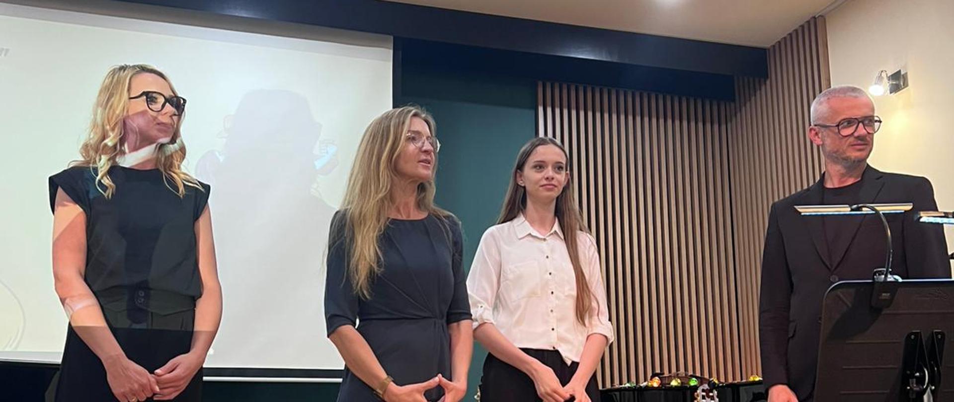 Na zdjęciu widać cztery osoby stojące na scenie. Trzy z nich są kobietami, a jedna mężczyzną. Kobiety mają różne stylizacje: jedna w ciemnej bluzce, druga w szarej sukience, a trzecia w białej bluzce. Mężczyzna jest ubrany w czarny strój. W tle znajduje się ekran oraz elementy sceny, w tym krzesła. Atmosfera wygląda na formalną, sugerując, że to może być zakończenie występu lub prezentacji.