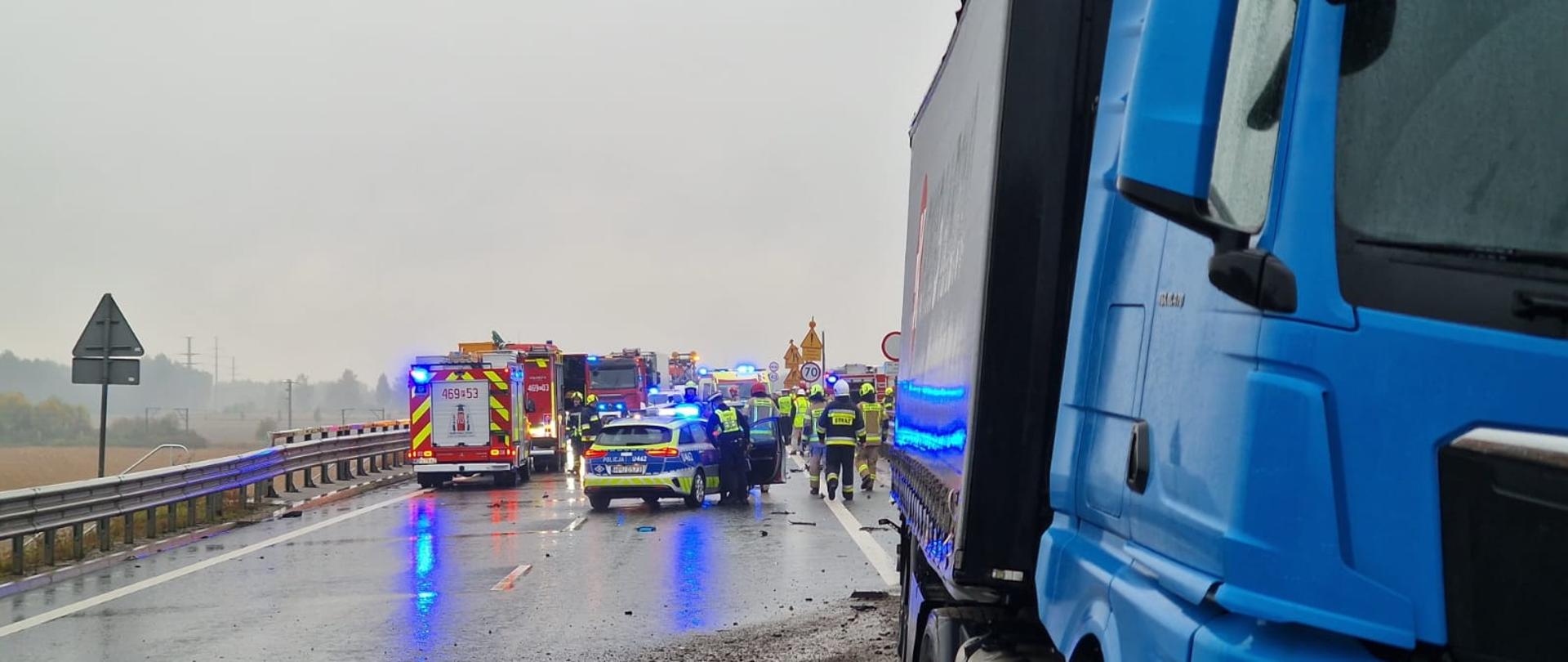 Zdjęcie przedstawia miejsce zdarzenia drogowego na mokrej trasie, sfilmowane z perspektywy niebieskiej ciężarówki marki MAN stojącej na prawym poboczu na pierwszym planie. Na nawierzchni wokół ciężarówki rozrzucone są drobne odłamki i kamienie. W głębi drogi, na zablokowanych pasach ruchu, widoczne są pojazdy służb ratowniczych z włączonymi błyskowymi światłami ostrzegawczymi: czerwone wozy strażackie, radiowóz policyjny oraz karetka pogotowia. Wokół nich pracują strażacy w ubraniach ochronnych i hełmach oraz policjanci. Po lewej stronie trasy ciągną się bariery ochronne, a w tle widać szare, zachmurzone niebo.
