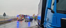 Zdjęcie przedstawia miejsce zdarzenia drogowego na mokrej trasie, sfilmowane z perspektywy niebieskiej ciężarówki marki MAN stojącej na prawym poboczu na pierwszym planie. Na nawierzchni wokół ciężarówki rozrzucone są drobne odłamki i kamienie. W głębi drogi, na zablokowanych pasach ruchu, widoczne są pojazdy służb ratowniczych z włączonymi błyskowymi światłami ostrzegawczymi: czerwone wozy strażackie, radiowóz policyjny oraz karetka pogotowia. Wokół nich pracują strażacy w ubraniach ochronnych i hełmach oraz policjanci. Po lewej stronie trasy ciągną się bariery ochronne, a w tle widać szare, zachmurzone niebo.
