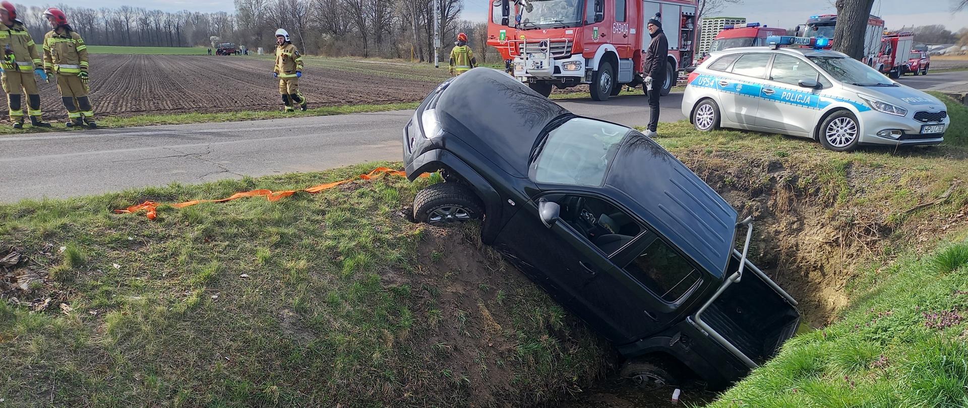 Na zdjęciu czarny samochód osobowy w rowie , w oddali samochód pożarniczy, radiowóz oraz strażacy
