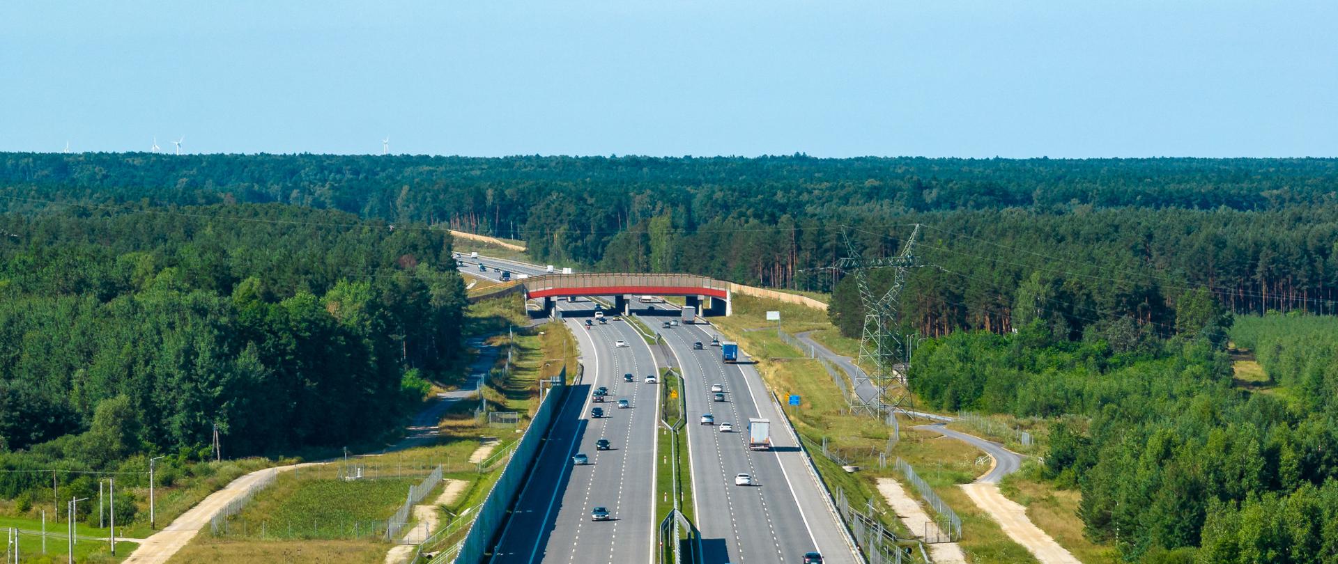Ruch pojazdów na autostradzie A1 w okolicach Radomska. Trzy pasy ruchu w każdą stronę. Środkowej części zdjęcia nad autostradą znajduje się przejście dla zwierząt. Po obu stronach drogi rośnie gęsty las.