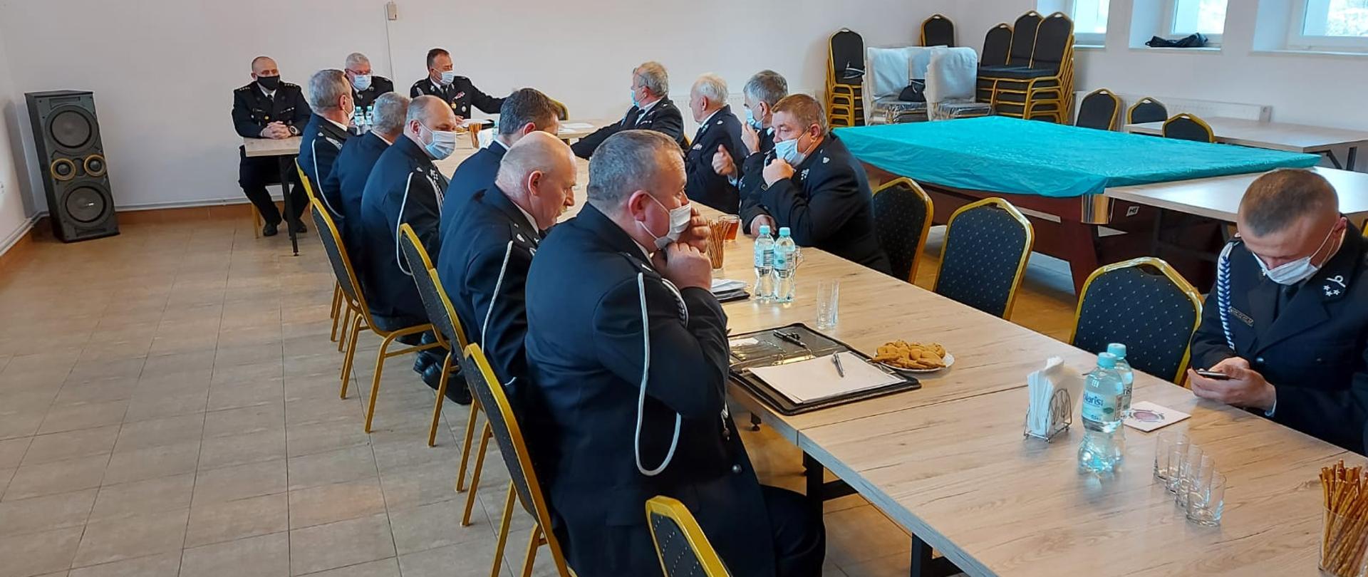 Na zdjęciu widać strażaków OSP w mundurach galowych którzy siedzą przy stole na którym znajdują się przekąski i woda do picia. Jeden stół na końcu ustawiony jest w poprzek do reszty, siedzą przy nim władze OSP oraz komendant powiatowy PSP w Ustrzykach Dolnych. Zdjęcie wykonane zostało na dużej sali. 