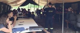 Zdajecie przedstawia strażaka z Jednostki Ratowniczo - Gaśniczej Dubiecko uczestniczącego w spotkaniu z dziećmi i młodzieżą. Szkolenie odbywa się w namiocie w tle widać las