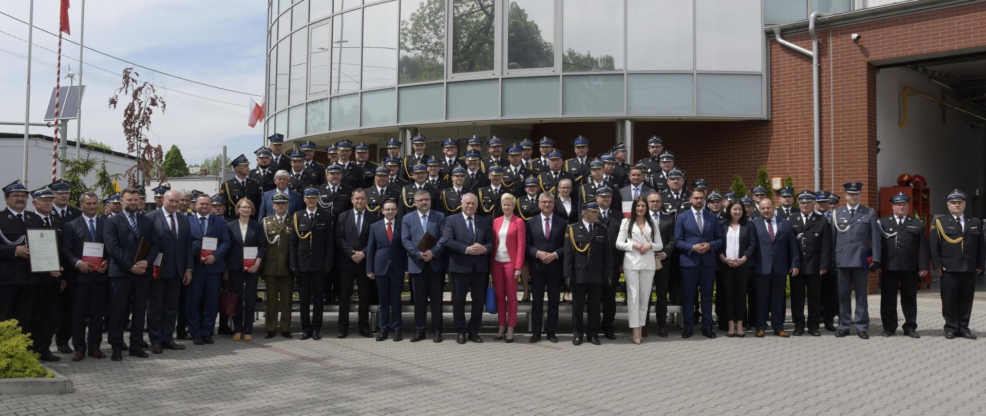 Na pierwszym planie widać stojących w szeregu uczestników uroczystości Powiatowych Obchodów Dnia Strażaka w dniu 13 maja 2022 m.in.: posłów, senatorów, przedstawicieli władz samorządowych, strażaków Państwowej i Ochotniczej Straży Pożarnej. W tle budynek strażnicy Komendy Powiatowej Państwowej Straży Pożarnej w Bochni