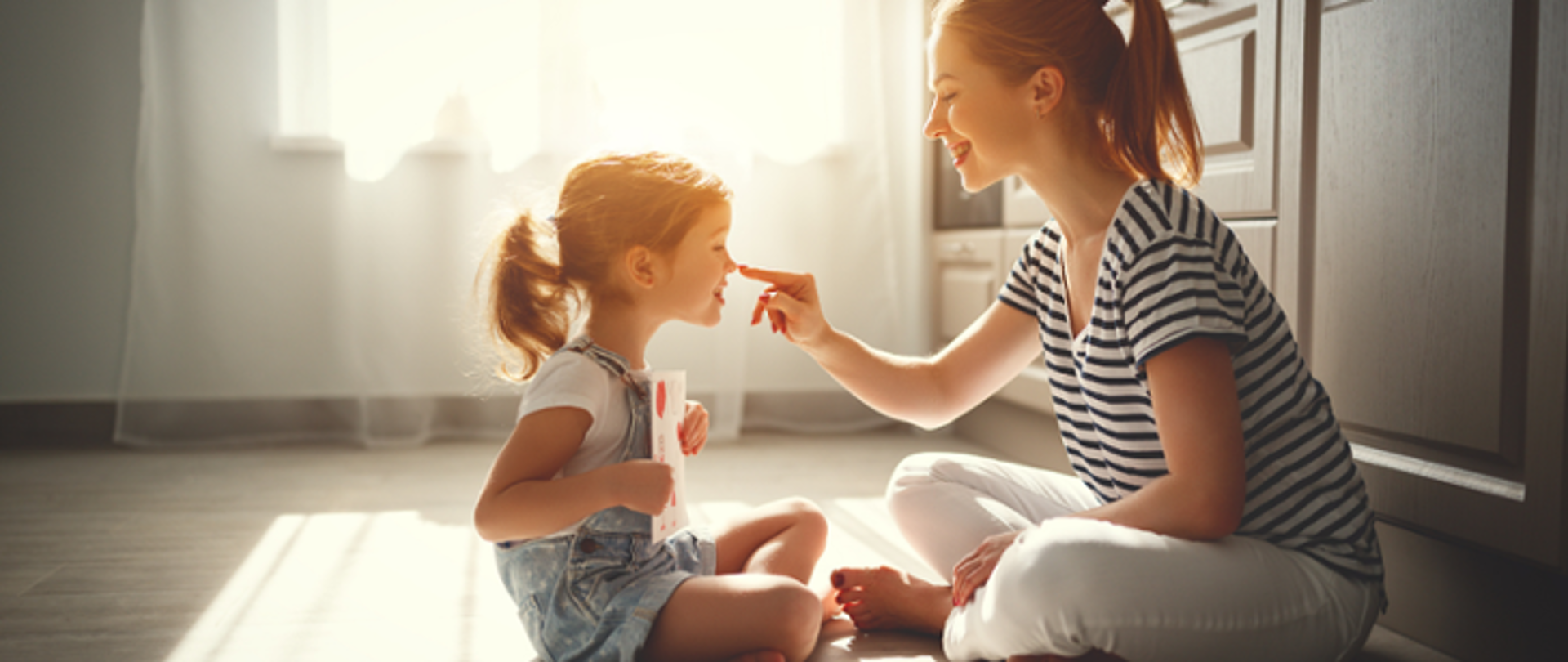 Na zdjęciu znajduje się dorosła kobieta wraz z małą dziewczynką. Obie widnieją w pozycjach siedzących. Są zwrócone ku sobie. W tle widoczne jest wnętrze pokoju.