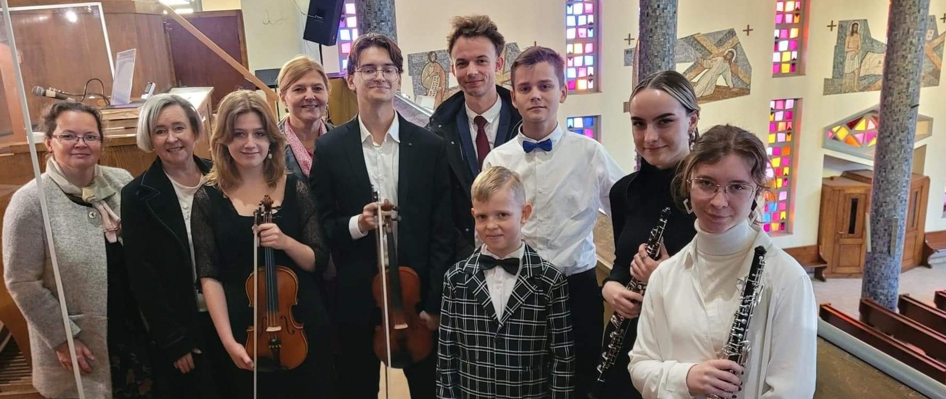 Na zdjęciu grupa młodzieży z nauczycielami. Wszyscy ubrani odświętnie. Dwie osoby trzymają w ręku skrzypce ze smyczkiem, a dwie klarnet. Zdjęcie wykonane we wnętrzu kościoła, na dole widać ławki dla wiernych oraz konfesjonał
