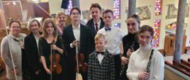 Na zdjęciu grupa młodzieży z nauczycielami. Wszyscy ubrani odświętnie. Dwie osoby trzymają w ręku skrzypce ze smyczkiem, a dwie klarnet. Zdjęcie wykonane we wnętrzu kościoła, na dole widać ławki dla wiernych oraz konfesjonał