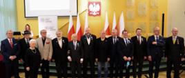 Na zdjęciu grupa osób odznaczonych Krzyżem Wolności i Solidarności. W sumie jest 11 osób. Za nimi widać kobietę, prowadzącą spotkanie. 