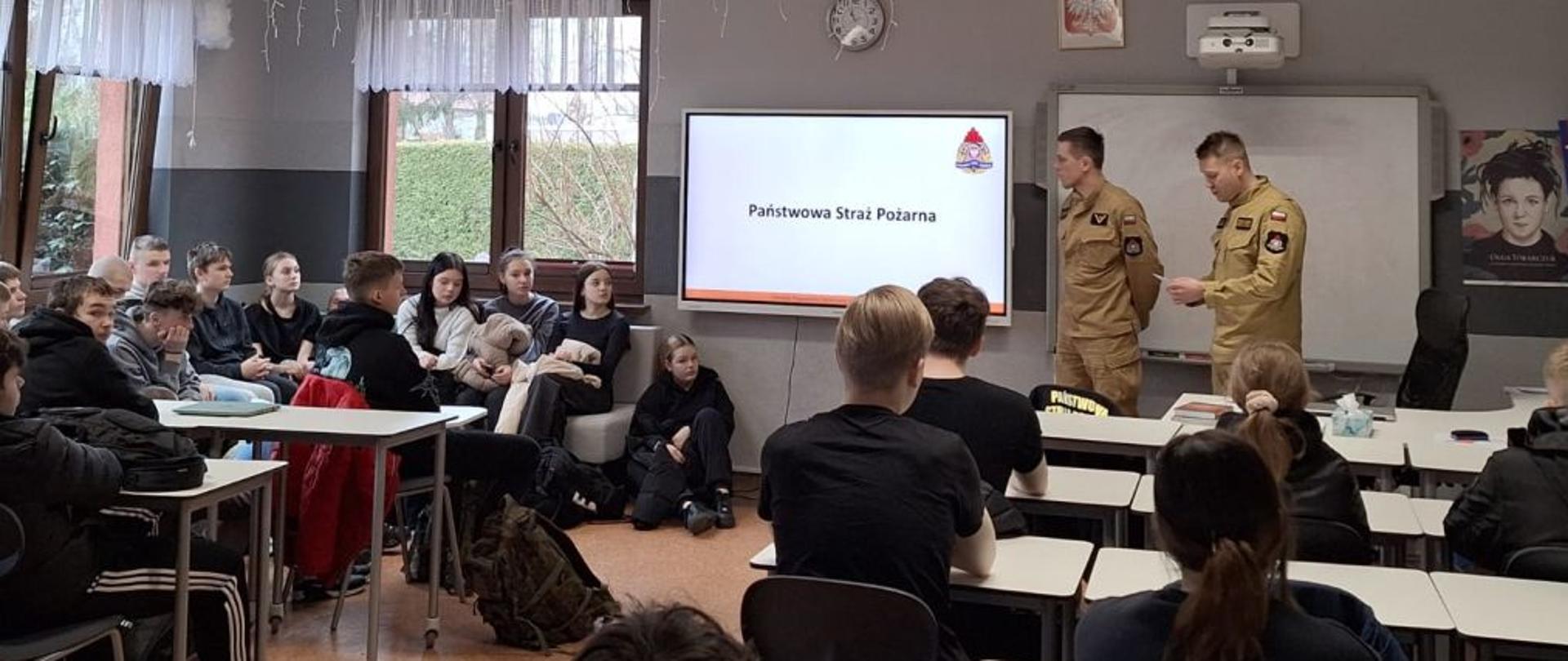 Szerszy kadr ukazuje całą klasę, wypełnioną uczniami zainteresowanymi tematyką PSP. Strażacy stoją na tle tablicy i ekranu z prezentacją, omawiając szczegóły swojej służby. Widać zaangażowanie zarówno prowadzących, jak i uczestników spotkania. Dekoracje sufitowe dodają sali wyjątkowego wyglądu. To ważne wydarzenie edukacyjne, mające na celu zainteresowanie młodych ludzi zawodem strażaka.