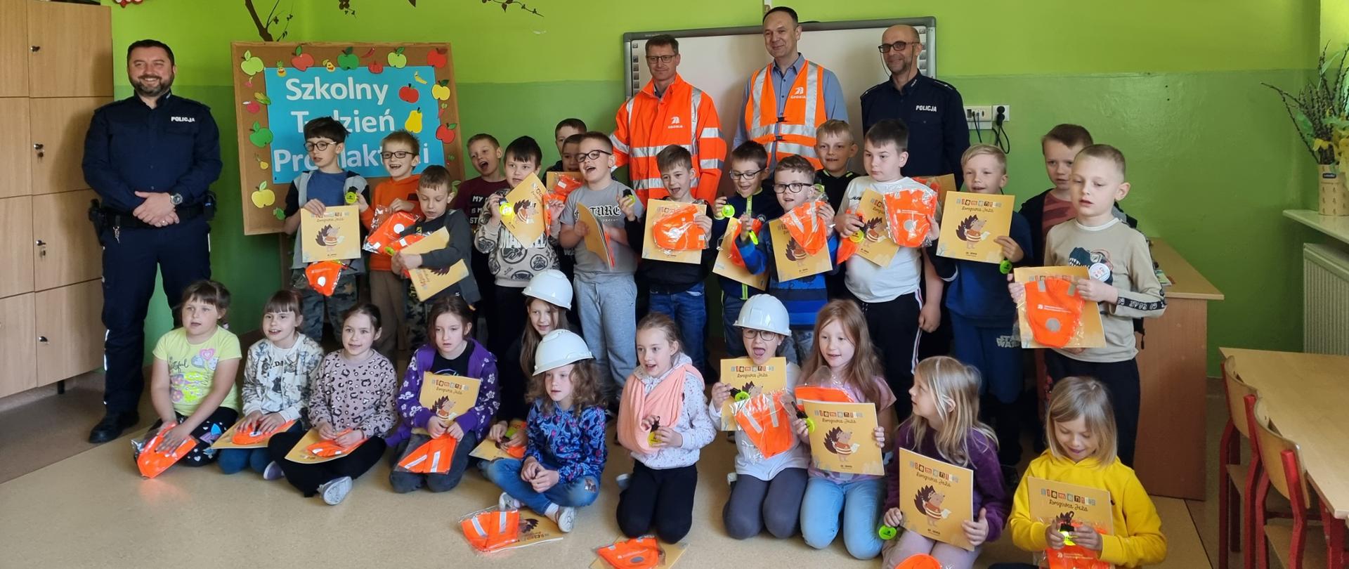 Lekcja bezpieczeństwa w Oblęgorku - w szkolnej sali dzieci trzymają elementy odblaskowe pomarańczowe i żółte, książeczki z drogowcem jeżem. Z tyłu dwóch policjantów w granatowych mundurach i dwóch drogowców w pomarańczowych odblaskowych ubraniach.
