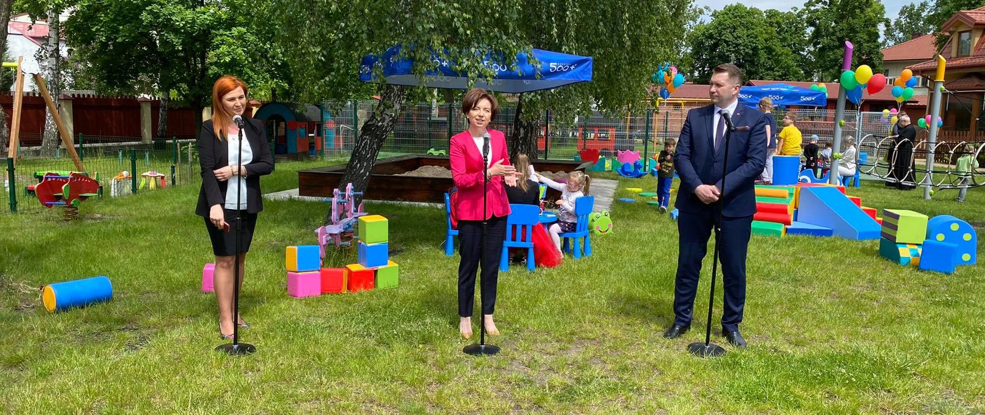 W ogródku zabaw stoją na zielonej trawie trzy osoby. Przed nimi mikrofony. W tle bawiące się dzieci.