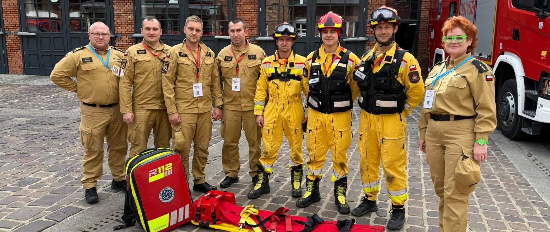 Na zdjęciu znajduje się grupa ośmiu strażaków (siedmiu mężczyzn i jedna kobieta) pozujących do pamiątkowej fotografii na placu przed remizą strażacką.
Ludzie i mundury:
Po lewej stronie i na prawym skraju stoją osoby w piaskowych (pustynnych/beżowych) mundurach służbowych Państwowej Straży Pożarnej.
W środku stoją trzej strażacy w specjalnych, żółtych kombinezonach bojowych (używanych np. do działań ratowniczych lub w poszukiwaniach), w kaskach/hełmach i specjalistycznym oporządzeniu.
Sprzęt ratowniczy:
Przez grupą, na brukowanym podłożu, leży żółto-czerwona deska ortopedyczna ze stabilizatorami i pasami do unieruchamiania poszkodowanego.
Obok stoi czerwono-żółta torba medyczna z napisem R112 i logo ratownictwa medycznego (Gwiazda Życia).
Tło:
W tle widać ceglany budynek strażnicy z numerowanymi bramami garażowymi (1, 2, 3).
Po prawej stronie zaparkowany jest czerwony wóz strażacki.
