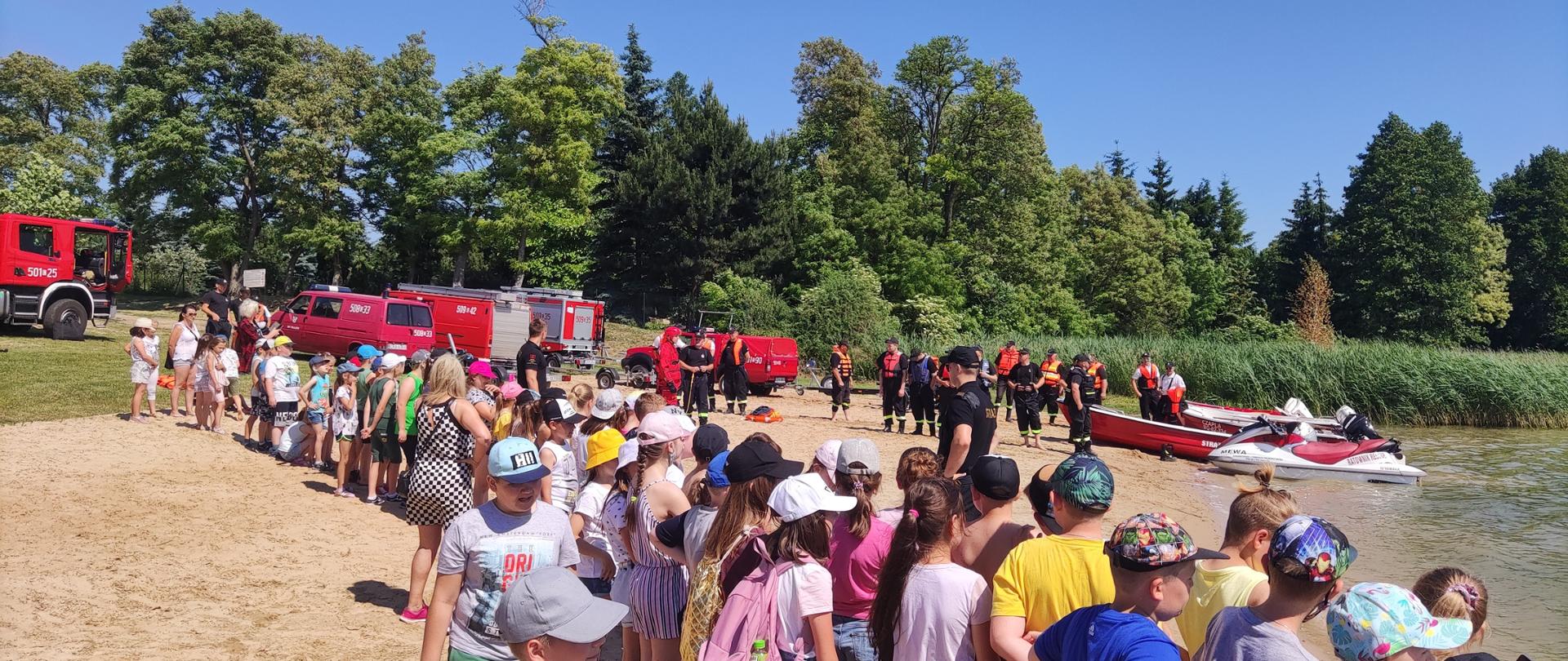 Zdjęcie przedstawia dzieci klas trzecich ze Szkół Podstawowych z Morzyc oraz z Radziejowa, strażaków PSP i OSP oraz sprzęt ratowniczy podczas pokazu ratownictwa wodnego.