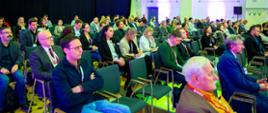 Osoby siedzą na krzesłach i słuchają przemówień podczas Konferencji Ewaluacyjnej