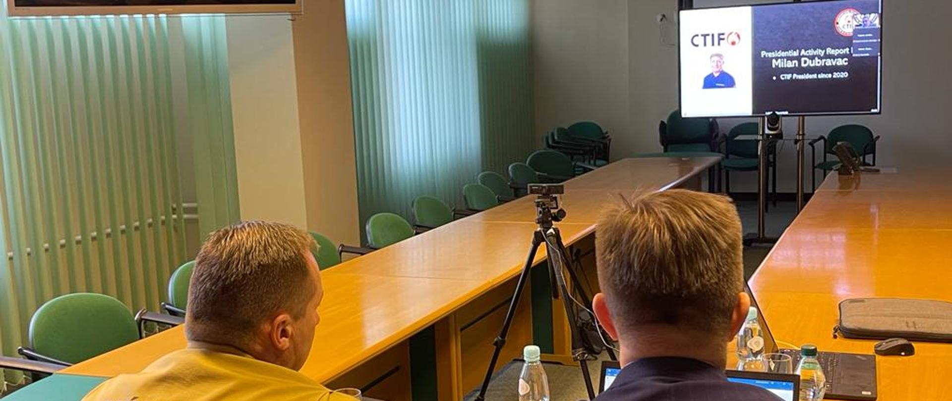 Zastępca Komendanta Głównego i Zastępca Dyrektora Biura Ochrony Ludności siedzą przy stole podczas udziału w formie wideokonferencji (widok z tyłu), przed nimi ekran z wyświetlonym slajdem