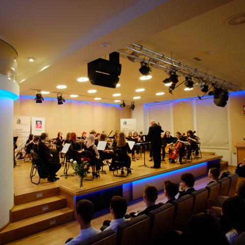 zdjęcie przedstawia orkiestrę szkoły, w ujęciu widoczne godło państwowe oraz płaskorzeźba z wizerunkiem patrona szkoły Feliksa Nowowiejskiego, w tle banery szkoły oraz miasta Szczecin