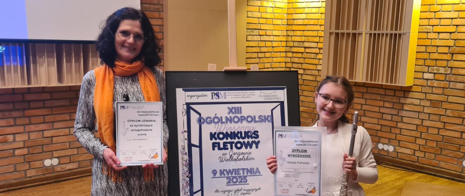 Nauczycielka i uczennica stojące przy plakacie z tytułem konkursu.