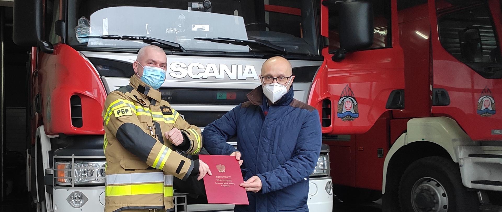 Zdjęcie przedstawia dwóch mężczyzn przekazujących sobie umowę darowizny w ozdobnej teczce. Mężczyzna z lewej strony kadru ma na sobie ubranie specjalne, natomiast mężczyzna z prawej strony kadru ma na sobie pikowaną kurtkę zimową. W tle znajdują się samochody strażackie. 