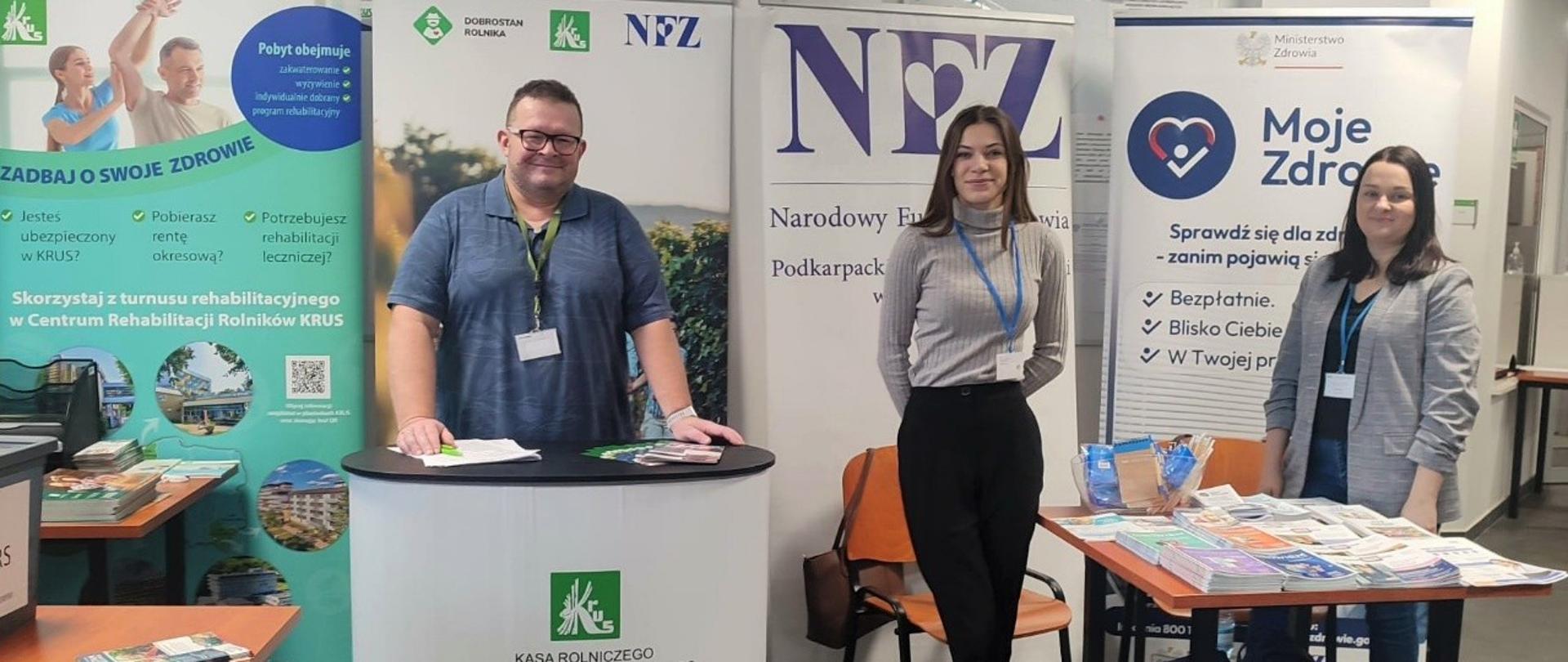 W sali obsługi interesantów znajduje się stoisko informacyjno-promocyjne Kasy oraz NFZ. Za stoiskami znajdują się roll-upy. Pracownik OR KRUS wraz z przedstawicielami NFZ pozują do zdjęcia.