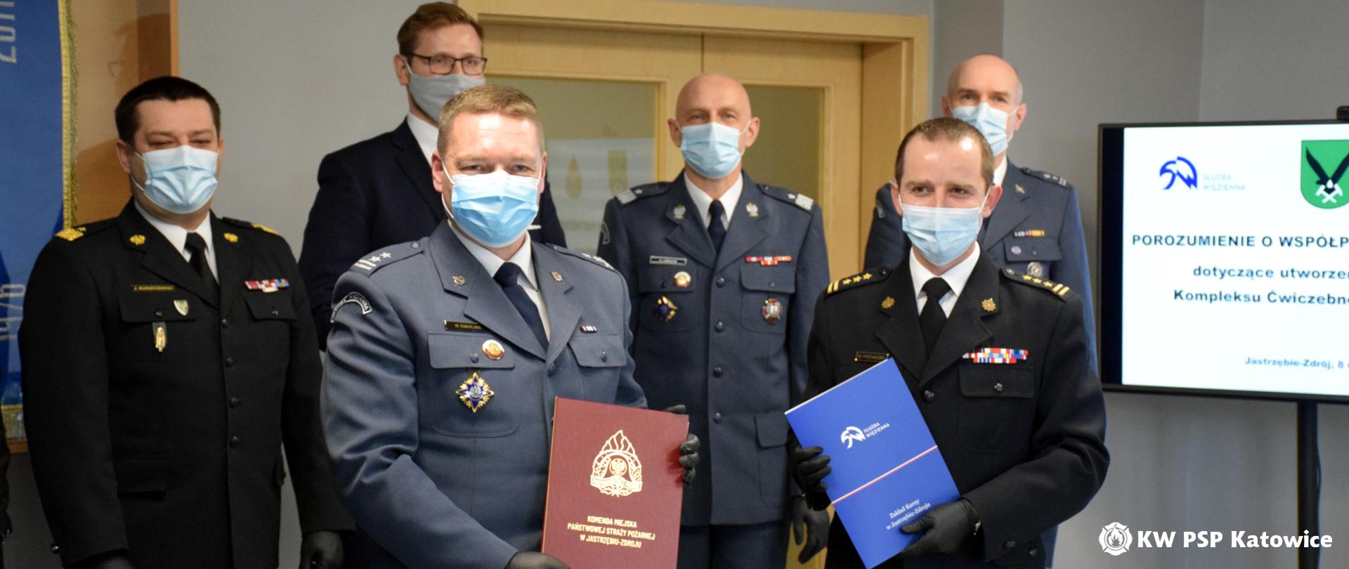 Na zdjęciu w sali Komendy Miejskiej PSP w Jastrzębiu Zdroju, na pierwszym planie widoczni są sygnatariusze porozumienia: komendant miejski PSP w Jastrzębiu-Zdroju st. bryg. Wojciech Piechaczek oraz dyrektor Zakładu Karnego w Jastrzębiu-Zdroju ppłk Wojciech Emerling. Ubrani w mundury i maseczki ochronne, trzymają w rękach podpisane porozumienie. W tle stoją zaproszeni goście : od lewej śląski komendant wojewódzki PSP nadbryg. Jacek Kleszczewski, sekretarza stanu w Ministerstwie Sprawiedliwości Pan Michał Woś, zastępca dyrektora generalnego Służby Więziennej gen. Andrzej Leńczuk oraz ppłk Łukasz Górny - dyrektor okręgowy Służby Więziennej w Katowicach.
