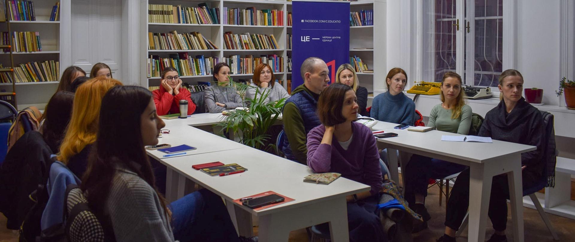W pomieszczeniu z regałami wypełnionymi książkami grupa osób siedzi przy stołach ustawionych w podkowę i patrzy się na prowadzącego zajęcia