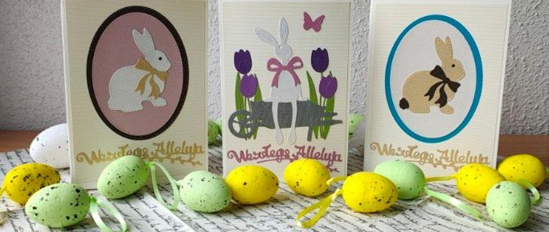 Trzy kolorowe kartki świąteczne ozdobione zajączkami i napisami "Wesołego Alleluja" stoją obok siebie na biało-czarnej serwetce. Na środkowej kartce siedzi zając wśród tulipanów. Na kartkach bocznych są zające naklejone na jajko. Przed karkami leżą jajeczka żółte i zielone