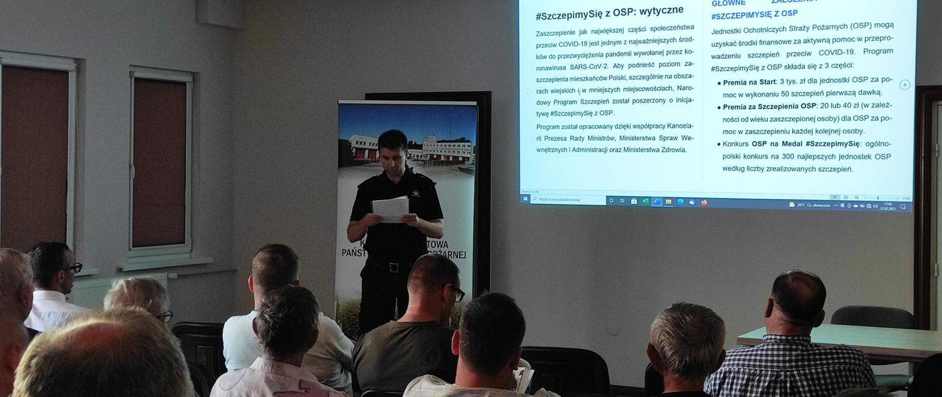 Na zdjęciu funkcjonariusz PSP w umundurowaniu dowódczo-sztabowym na tle baneru KP PSP w Tucholi. Na ścianie wyświetlane są slajdy prezentacji omawianego programu #SzczepimySię z OSP. Miejsce spotkania świetlica KPPSP w Tucholi. Na pierwszym planie druhowie z OSP.