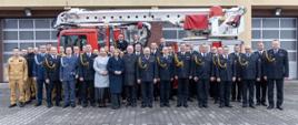 Zdjęcie grupowe uczestników uroczystości przed wozem strażackim PMT-25D - podnośnik hydrauliczny z koszem. Wśród obecnych funkcjonariusze PSP w galowych i bojowych mundurach oraz przedstawiciele lokalnych samorządów.