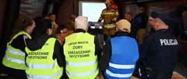 Uczestnicy ćwiczeń siedzą w namiocie na ławce. Jeden strażak mówi do nich. Na kamizelkach widnieją napisy Urząd Miasta Żory Zarzadzanie Kryzysowe. W tle widoczna prezentacja multimedialna. 