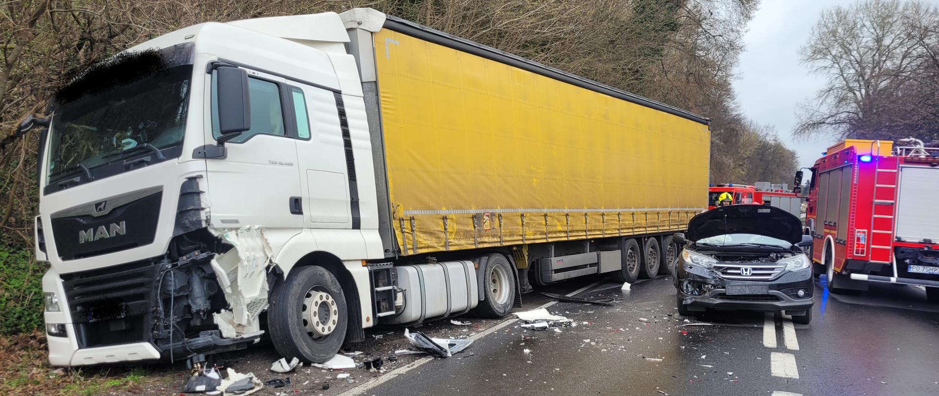  Rozbity pojazd osobowy, po lewej stronie naczepa ciężarówki, a w tle samochód pożarniczy