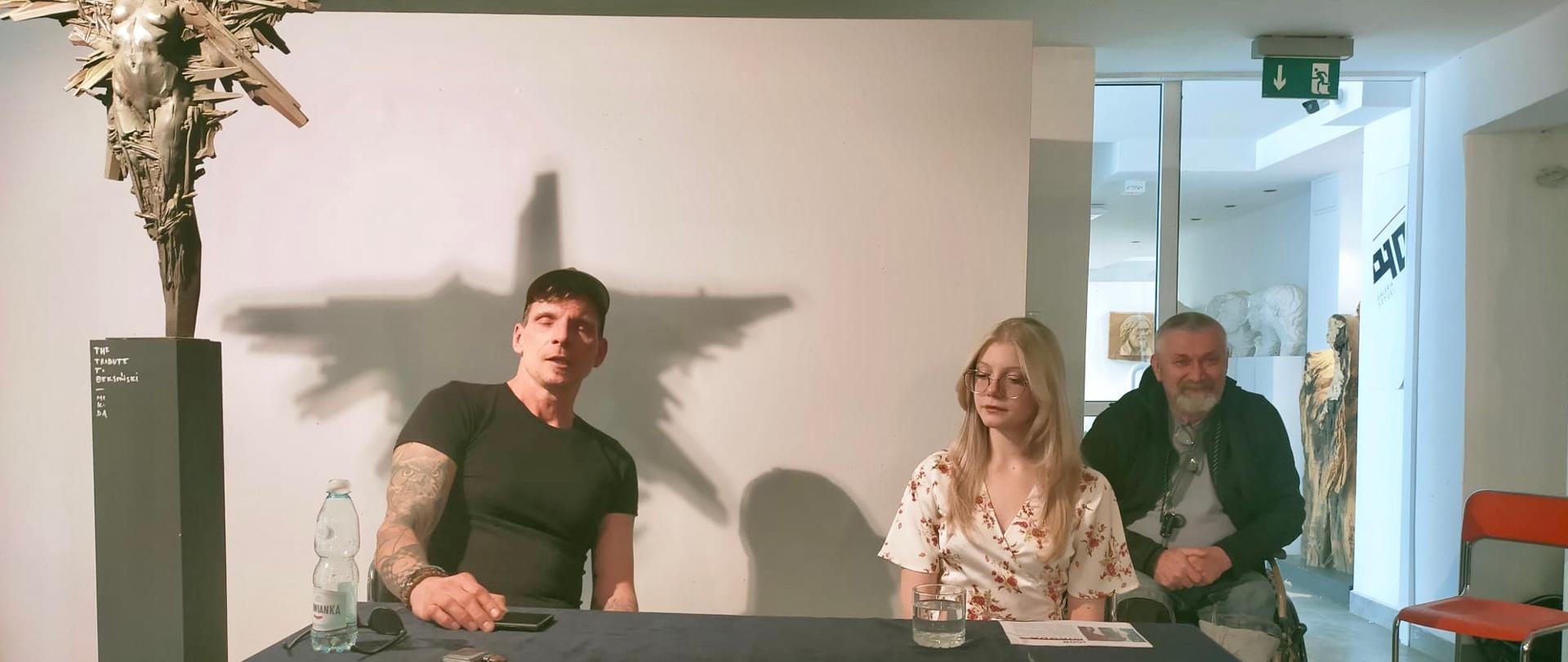 Zdjęcie przedstawia spotkanie autorskie odbywające się w nowoczesnym wnętrzu galerii sztuki.
Główne elementy:
Postacie: Za stołem przykrytym czarnym obrusem siedzą trzy osoby: po lewej mężczyzna w czarnym t-shircie z tatuażami, w środku młoda kobieta w jasnej sukience w kwiaty, a po prawej starszy, uśmiechnięty mężczyzna z brodą.
Rzeźba: Po lewej stronie stoi smukła, dynamiczna rzeźba o nieregularnych kształtach, zamontowana na wysokim postumencie. Rzuca ona na ścianę za siedzącymi osobami duży, wyraźny cień przypominający skrzydła lub postać w locie.
Tło: Jasne ściany galerii, w oddali widoczne przejście do innego pomieszczenia z kolejnymi dziełami sztuki oraz zielony znak wyjścia ewakuacyjnego.
Scena ma charakter kameralnej dyskusji lub wernisażu.