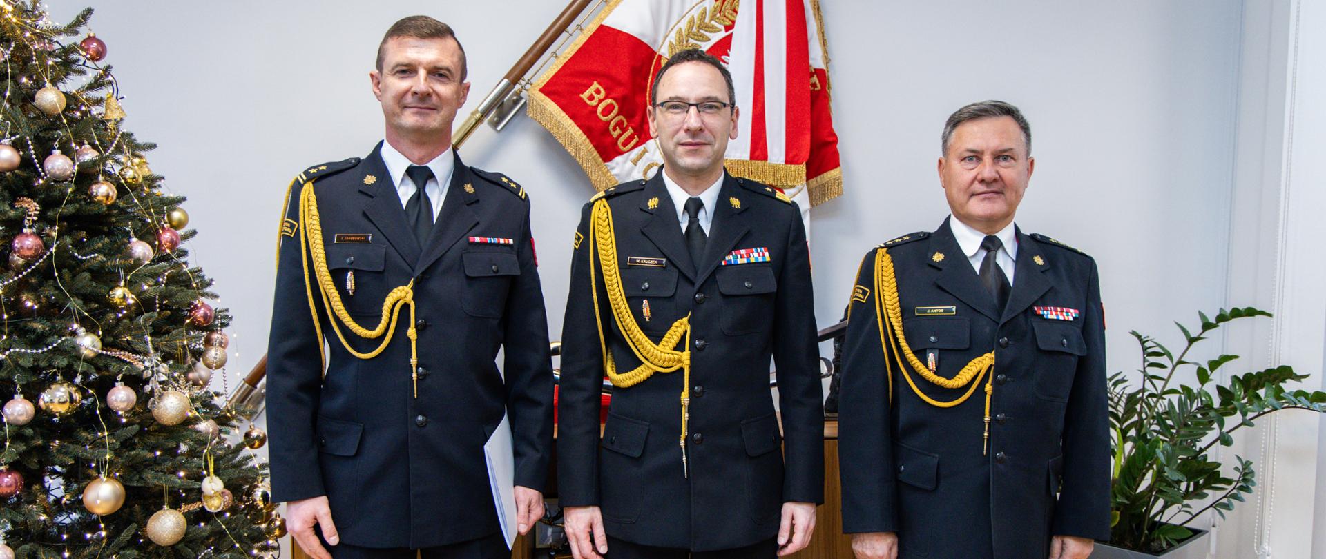 Trzech funkcjonariuszy PSP w galowych mundurach pozuje do zdjęcia w gabinecie; osoba po lewej trzyma białą teczkę. Po lewej stronie stoi ozdobiona choinka, a za nimi widoczny jest sztandar w barwach biało-czerwonych; obok znajduje się szafka i roślina doniczkowa.