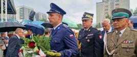 Policjant trzyma wiązankę, za nim stoją pomorski komendant wojewódzki Państwowej Straży Pożarnej przedstawiciel wojska oraz zaproszeni goście.