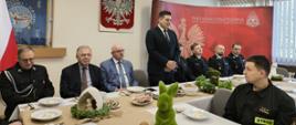 Przy stołach na których znajdują się dekoracje świąteczne i talerze, siedzą strażacy i zaproszenie goście. Strażacy ubrani są w mundury galowe zaproszeni goście odświętnie w garniturach. Jeden z zaproszonych gości w postawie stojącej przemawia. W tle logo Państwowej Straży Pożarnej oraz godło Polski i flagi