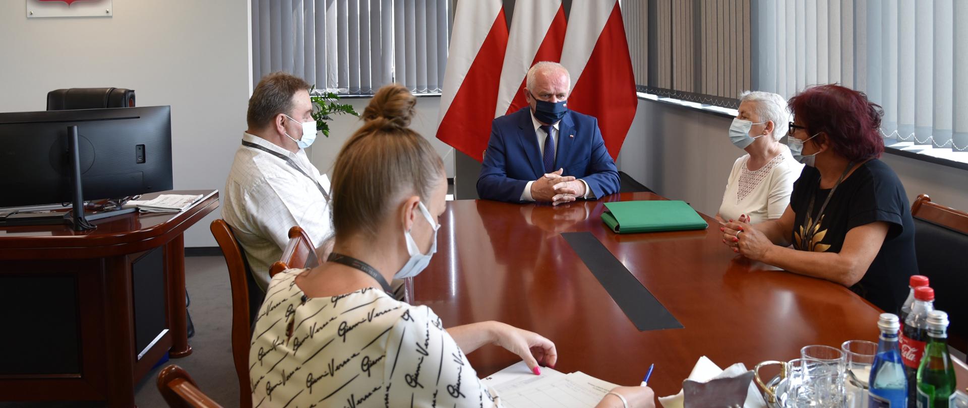 Wojewoda siedzi u szczytu stołu w swoim gabinecie. Przy stole, po obu stronach siedzą mieszkańcy i pracownica urzędu. W tle biało-czerwone flagi i biurko wojewody. 