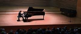 Mężczyzna gra na fortepianie na estradzie sali koncertowej, z przodu przed estradą widać od tyłu fragment widowni zapełnionej publicznością , nad estradą na widowni siedzi grupa młodzieży.