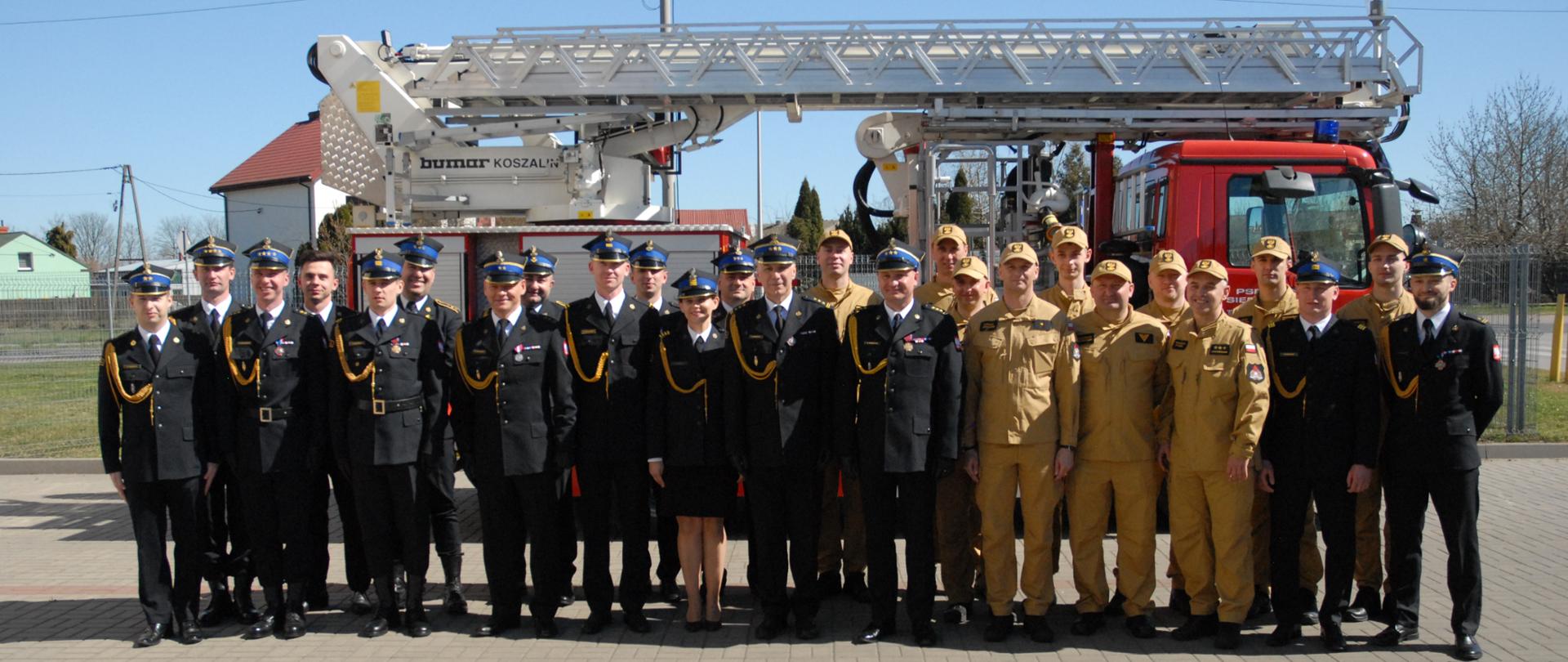 Zdjęcie grupowe strażaków w mundurach galowych i służbowych stojących na tle boku samochodu z drabiną mechaniczną. Zdjęcie wykonane w słoneczną pogodę. Pojazd i strażacy stoją na placu utwardzonym kostką betonową. 