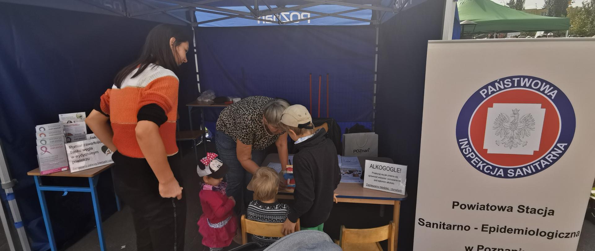 Na stoisku festynowym grupa dzieci uczestniczy w pokazie właściwego szczotkowania zębów, po prawej namiotu stronie stoi baner z logo Państwowej Inspekcji Sanitarnej
