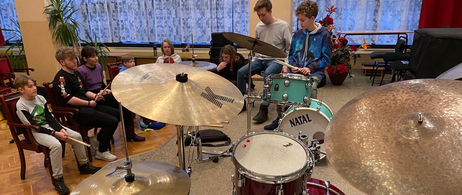 Kacper Kałuża z uczniem grający na scenie auli PSM na zestawie perkusyjnym, w tle sześciu uczniów siedzących na widowni
