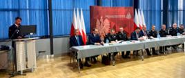 Mężczyzna w mundurze strażackim przemawiający przy mównicy. Obok mężczyźni siedzący przy stoliku prezydialnym. Za nimi ścianka PSP oraz flagi narodowe