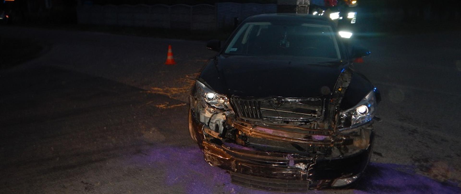 Zdjęcie przedstawia samochód koloru czarnego marki Skoda Octawia, uszkodzony z przodu, po zderzeniu z samochodem osobowym marki Nissan Primery.