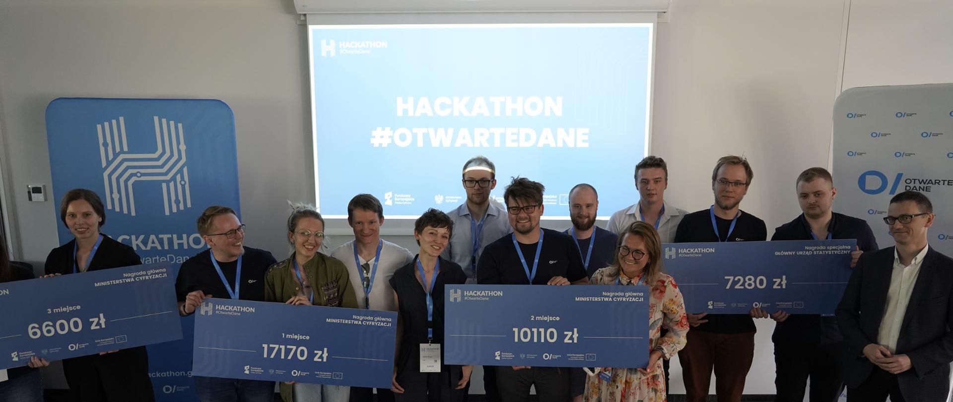 Zdjęcie grupowe organizatorów i laureatów hackathonu Otwarte Dane. Kilkanaście uśmiechniętych osób na tle ekranu i rollupów z logotypami wydarzenia. Różne osoby trzymają tabliczki z informacjami o zdobytych nagrodach (które miejsce i jaka kwota pieniędzy). Tabliczki dotyczą 1,2 i 3 miejsca oraz nagrody specjalnej.